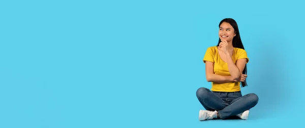 黄色のTシャツの思考で思慮深い陽気な魅力的な若い中国の女性は ハスの位置に座って 空のスペースを見て 青の背景 スタジオに孤立した 広告やオファー 人間の感情や販売 — ストック写真