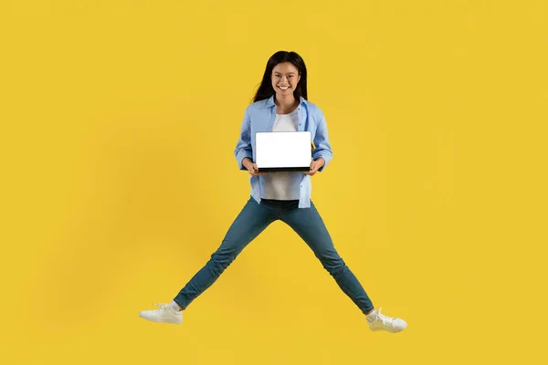 Smiling young chinese woman student in casual jumping, showing laptop with blank screen, isolated on yellow background, studio. Device for study and education, ad and offer, huge sale, online shopping