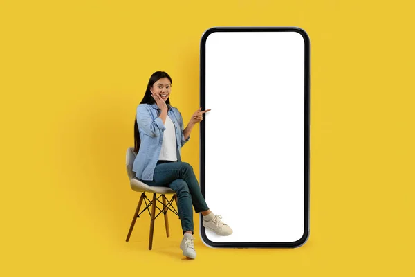 微笑震惊地坐在椅子上的年轻的中国女学生 手指在巨大的智能手机与空屏幕 孤立在黄色的背景下 工作室 应用程序 广告和提供 销售和好消息 — 图库照片