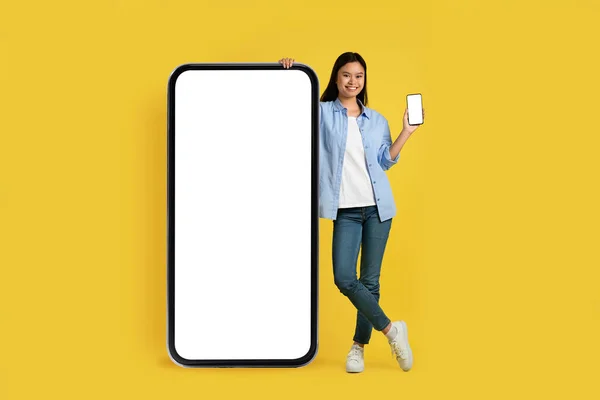 New app, digital and ad offer. Smiling young japanese lady student in casual shows phone, stands near huge smartphone with empty screen, isolated on yellow background. Gadget for study and shopping