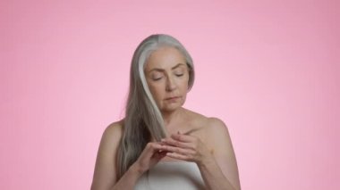 Yaşlanma ve saç sorunları. Yaşlı, gri saçlı, kuru saçlarına bakan, çatık kaşlı, üzgün, pembe arka planda, yavaş çekimde üzgün bir kadının stüdyo resmi.