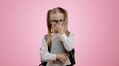 Küçük dahi. Sırt çantası ve kitaplar taşıyan, gözlüğünü takan, kameraya gülümseyen, pembe arka planda, ağır çekimde