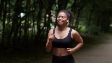 Aktif yaşam tarzı konsepti. Genç Afro-Amerikan bayan sporcu parktaki sabah koşusundan keyif alıyor, kablosuz kulaklıkla müzik dinliyor, yavaş çekim takip ediyor.