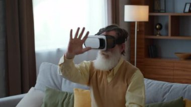 Yaşlılar için gelecek teknolojileri. Büyükbabanın VR kulaklık taktığı yakın plan portresi evde artırılmış gerçekliğin tadını çıkarıyor, görünmez düğmelere basıyor, yavaş çekim yapıyor, takip ediyor.