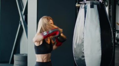 Dişi kickboks. Aktif motive olmuş boks eldivenli kadın sporcu spor salonunda kum torbasını yumrukluyor, spor salonunda kendini savunma pratiği yapıyor, yavaş çekim, serbest alan