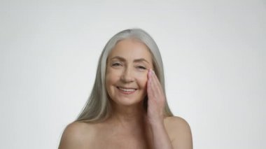 Yaşlanma karşıtı yüz yogası. Güzel yaşlı gri saçlı bayan kırışıklık önleyici yüz masajı yapıyor, kameraya gülümsüyor, beyaz stüdyo arka planı, yavaş çekim.