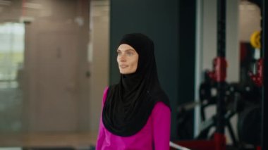 Geleneksel tesettürlü, orta yaşlı Müslüman kadın spor salonunda çalışıyor, dambılları kaldırıyor, kaslarını çalıştırıyor, ağır çekimde çalışıyor.