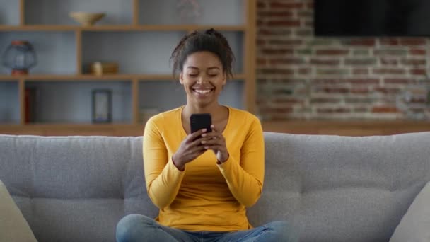 面白いインターネットコンテンツ 幸せなアフリカ系アメリカ人女性は スマートフォンでオンラインニュースを読んで笑い ユーモアを楽しみ 自宅でソファに休んで スローモーション フリースペース — ストック動画