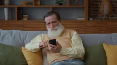 Yaşlılar için sosyal medya. Modern yaşlı sakallı adamın kapalı bir portresi. İnternet 'te sörf yapıyor, internetten haberleri okuyor, evdeki koltukta oturuyor, yavaş çekim, serbest alan.
