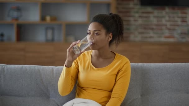 日常生活中的健康习惯 非洲裔美国女人用玻璃杯喝水 坐在沙发上 跟踪镜头 慢动作 自由自在的形象 — 图库视频影像
