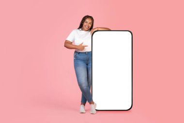Pembe arka planda, stüdyoda kameraya gülümseyen boş ekranlı, geniş akıllı telefonu işaret eden Afrikalı Amerikalı aşırı kilolu kadın. Bu Mobil Uygulamayı Kontrol Et. Model, Tam Uzunluk