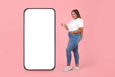Siyah kilolu kadın, Pembe Stüdyo Arkaplanında Ayakta Duran Boş Ekran Pozu Veren Büyük Telefonun Yanındaydı. Neşeli Kadın Reklamcılığı Büyük Mobil Teklif. Tam Uzunluk, Model