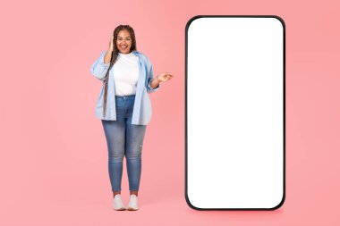 Pembe Stüdyo 'nun arka planında duran boş ekranlı büyük telefonu işaret eden mutlu siyah kadın. Dişi Önerisi Yeni Mobil Uygulama. Model, Tam Uzunluk