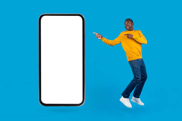 Mobile app concept. Cheerful african american guy dancing by big cell phone over blue studio background, african american man pointing at smartphone with white empty screen, mockup, full length shot