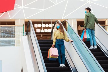 Hafta sonu alışveriş merkezinde. Modern süpermarkette, hareketli merdivenlerde duran insanlar koruyucu yüz maskeleri takıyorlar. Erkek ve Kadın Alıcılar Hipermarkette Zaman Geçiriyorlar