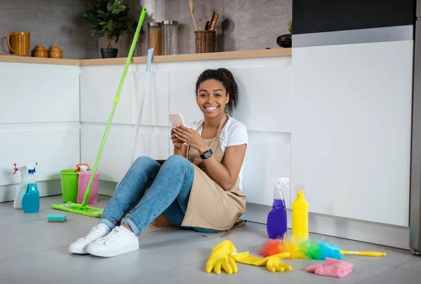快乐的年轻黑人女士坐在地板上 带着清洁用品 一边聊天 一边用电话在厨房里冲浪 业余时间放弃家务活 家庭清洁和卫生应用软件 — 图库照片