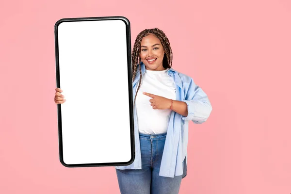 Happy African American Lady Holding Large Cellphone With Blank Screen Advertising Mobile App Standing Over Pink Studio Background, Smiling To Camera. Great Application. Mockup