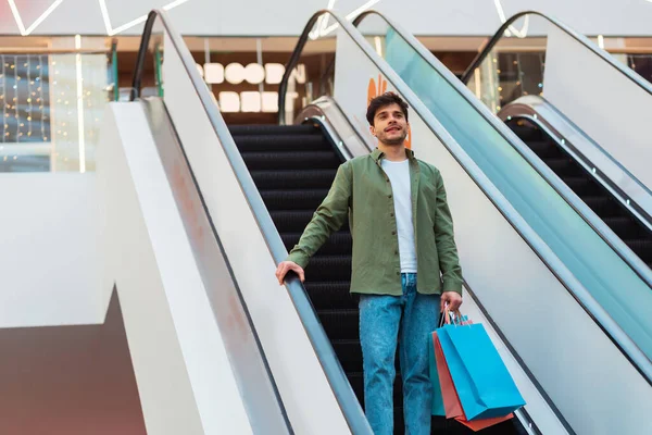 Shopping And Sales. Man Holding Shopper Bags Standing On Moving Stairs In Modern Mall Indoors. Millennial Male Buying New Clothes During Great Sales Season