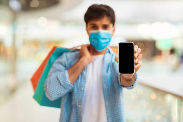 Mobile Shopping App. Man Showing Phone With Empty Screen Holding Shopper Bags Posing Wearing Face Mask Standing In Mall Indoors. Selective Focus On Smartphone