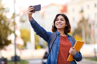 Neşeli Arap kız öğrenci dışarıda akıllı telefonuyla selfie çekiyor, çalışma kitaplarını tutan mutlu genç Orta Doğulu kadının portresi ve dersten sonra cep telefonuyla fotoğraf çekiyor, Boşluğu Kopyala