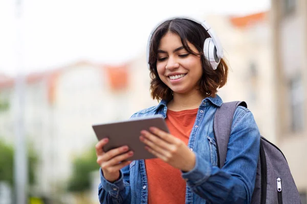 Smiling Middle Eastern Female Student In Wireless Headphones Using Digital Tablet Outdoors, Happy Young Arab Woman Browsing Internet On Modern Gadget While Walking After Classes, Copy Space