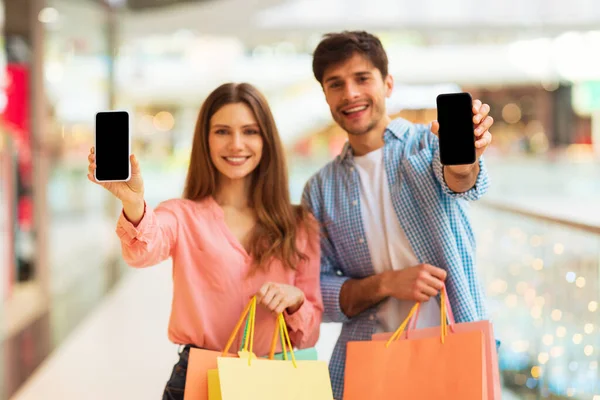 Mobile Shopping Application. Spouses Showing Cellphones With Empty Screens For App Advertisement Standing In Modern Mall Indoor, Smiling To Camera. Mockup, Shallow Depth