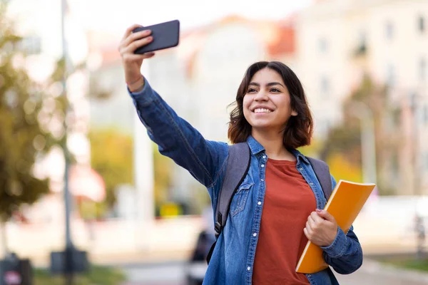 Cheerful Arab Female Student Taking Selfie With Smartphone Outdoors, Portrait Of Happy Young Middle Eastern Woman Holding Workbooks And Capturing Photo With Mobile Phone After Classes, Copy Space