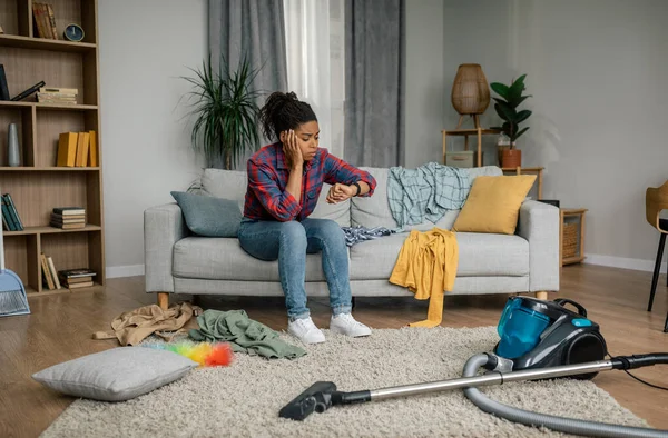 クリーニングの助けが必要です 掃除機付きの悲しい疲れた若い黒人女性はリビングルームのインテリアで時計を見ています 家庭だけで家事の多くは 混乱や障害 ストレス ライフスタイルや感情 — ストック写真