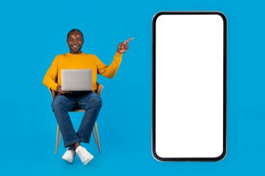 Mavi stüdyo arka planında gündelik koltukta oturan mutlu orta yaşlı siyah adam, modern bilgisayar kullanıyor, beyaz ekranlı büyük cep telefonunu işaret ediyor, maketini yapıyor. İş gezici uygulama kavramı