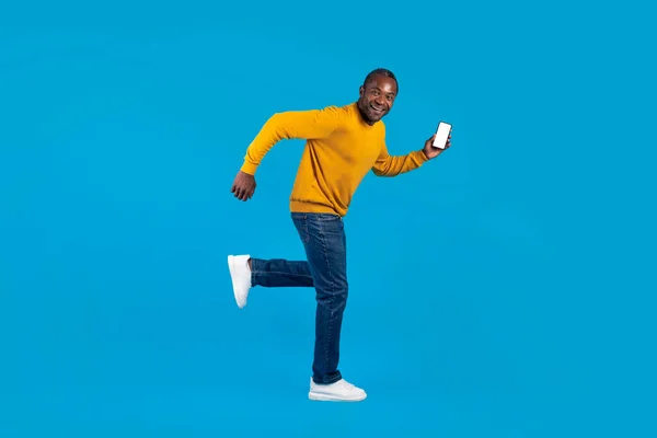 Funny attractive black man in casual outfit running away with modern smartphone with white blank screen in his hand, blue studio background, side view, copy space, full length shot, mockup