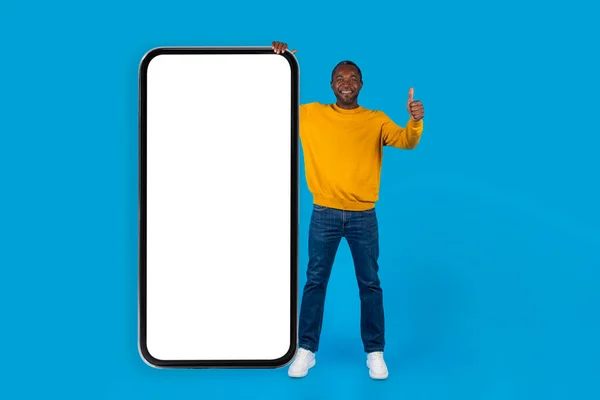 快乐的中年非洲男人 随意地站在巨大的智能手机旁 白色的空白屏幕 露出大拇指和微笑 蓝色的工作室背景 复制的空间 — 图库照片