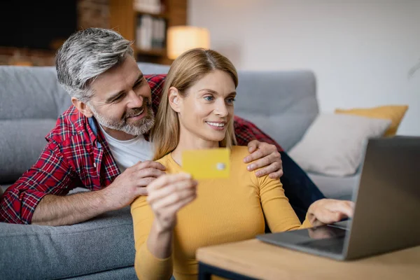 笑的欧洲中年男人抱着女人用信用卡 在笔记本电脑上打字 在客厅里网上购物 银行设备 购物狂销售 广告和提供适当的Covid 19病毒 — 图库照片