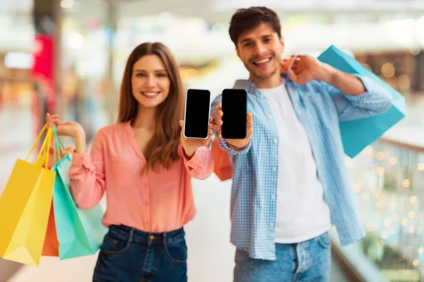 現代のハイパーマーケットでカラフルなショッパーバッグとモバイルショッピングアプリケーションのポーズをお勧めスマートフォン画面を示す陽気なカップル 携帯電話に焦点を当てる モックアップ — ストック写真