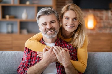 Aşk ve ilişki. Orta yaşlı mutlu Avrupalı eş kocasını kucaklıyor, rahat ve romantik bir şekilde oturma odasının iç kısmında kanepede oturuyor. Boş zaman, insan duyguları, dinlenme ve rahatlama.