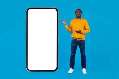 Neşeli orta yaşlı, günlük kıyafetli, mavi stüdyo arka planında tek başına poz veren, beyaz ekranlı büyük bir cep telefonunu gösteren, güzel bir online teklif sunan, model, tam boy