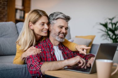 Neşeli, olgun Avrupalı eş kocaya sarılıyor, bilgisayara bakıyor, rahat bir oturma odasında oyun oynuyor. Cihazla eğlence, online alışveriş, covid-19 salgını sırasında sosyal medya sohbeti