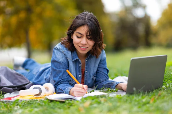 远程学习 年轻的阿拉伯女性 带着笔记本电脑准备参加户外考试 微笑着参加在线课程 使用电脑 躺在公园草坪上记笔记 — 图库照片