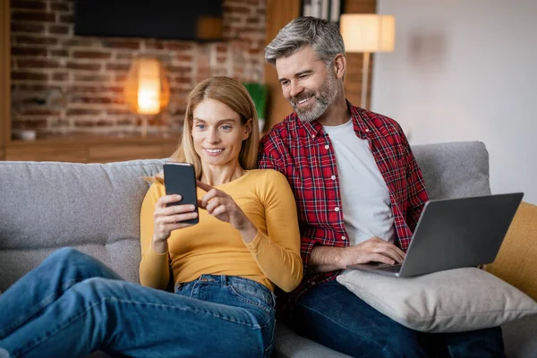 快乐成熟的欧洲男人 带着笔记本电脑看智能手机 妻子在社交网络上聊天 在客厅的沙发上享受自由时间 用于工作和上网 设备成瘾的新应用程序 — 图库照片