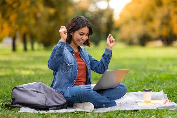 好消息 快乐的阿拉伯女学生与笔记本电脑户外庆祝成功 兴奋的中东年轻妇女与计算机坐在秋天公园的毛毯上 挥动拳头 复制空间 — 图库照片