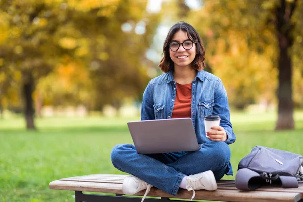 市内のベンチに座って遠隔学習を楽しみながら ノートパソコンを使ってコーヒーを飲みながら中東の若い女性を笑顔にする 陽気なアラブ女性学生 — ストック写真