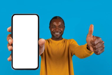 Pozitif yakışıklı orta yaşlı Afro-Amerikan adam beyaz ekranlı ve başparmağıyla yepyeni bir akıllı telefon gösteriyor, en yeni mobil uygulamayı tavsiye ediyor, mavi stüdyo arka planı, yakın plan,