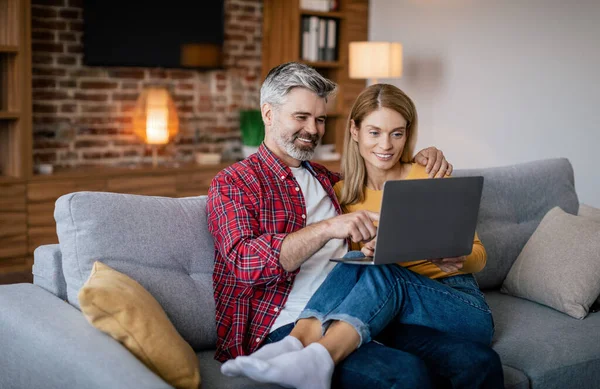 Glad mature european couple hug, look at laptop, surfing in internet, enjoy free time, sit on sofa in living room interior. Video call, meeting remotely, communication, ad and offer during covid-19