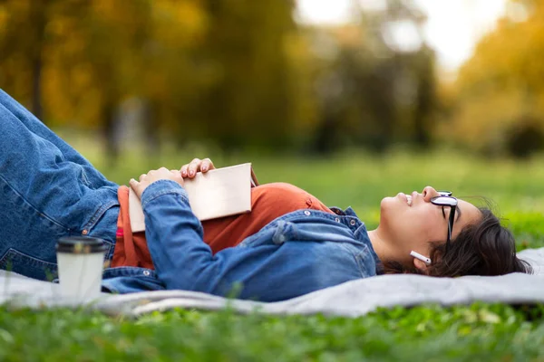 Açık hava eğlencesi. Gözlüklü inek genç bir Arap kadın kitapla çimlerde uzanıyor, Milenyum Orta Doğu 'lu bir kadın parkta dinleniyor ve kulaklıklarla müzik dinliyor, yan görüş