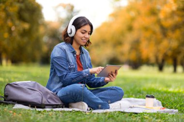 Dışarıda. Rahat ol. Sonbahar parkında battaniyenin üzerinde otururken dijital tablet kullanan, kablosuz kulaklık takan gülümseyen genç Arap kadın, modern Gadget ile çimlerin üzerinde dinlenen mutlu Orta Doğulu kadın.