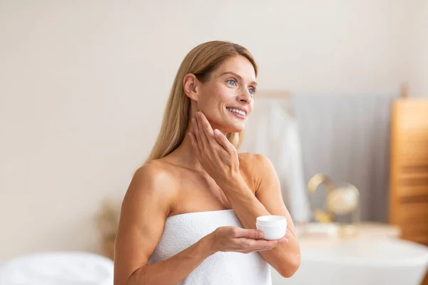 日常护肤 漂亮的中年女性用乳霜和保湿的罐子 裹着毛巾站在浴室的内部 — 图库照片