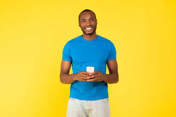 明るい黒の男は スマートフォンのウェブサーフィンを保持し 黄色のスタジオの背景に立ってテキストポーズ カメラに笑みを浮かべて 素晴らしいアプリケーションとモバイルオファーのコンセプト — ストック写真