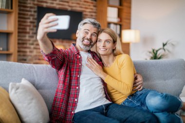 Neşeli olgun Avrupalı erkek ve karısı uzaktan video görüşmesi yapıyor, sosyal ağlar için fotoğraf çekiyor, sıcak oturma odasındaki kanepede oturuyor. Modern cihaz, toplantı uygulaması ve covid-19 salgını nedeniyle blog