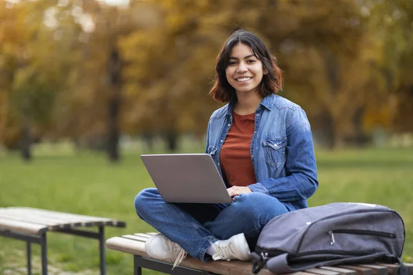 市内公園の外で試験のために準備しているコンピュータでベンチの屋外に座ってラップでラップトップで笑顔の若い中東女性学生の肖像画 コピースペース — ストック写真