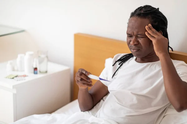 生病的年轻非洲裔美国人坐在床上时拿着温度计 生病的黑人男人检查体温和发情头 忍受季节性流感症状 复制空间 — 图库照片