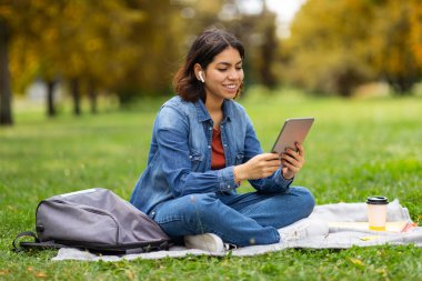 Genç gülümseyen Arap kadın dijital tabletle parkta dinleniyor, Milenyum Orta Doğu 'lu kadın çimlerin üzerinde oturuyor ve modern Gadget kullanıyor, kablosuz kulaklık takıyor, uzay kopyası yapıyor.